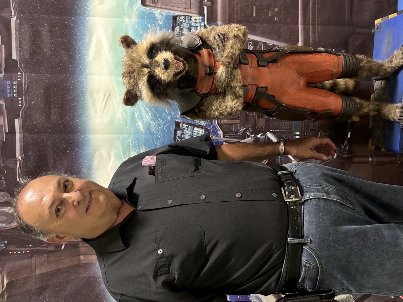 Russ Drew smiling beside a costumed raccoon character figure in front of a space-themed backdrop